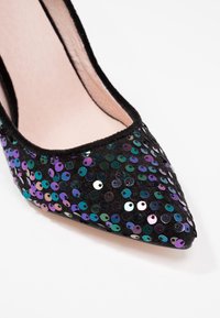 Chaussure noire pointue avec des sequins irisés violets, verts et bleus à l'avant et une doublure intérieure rose douce sur fond blanc.