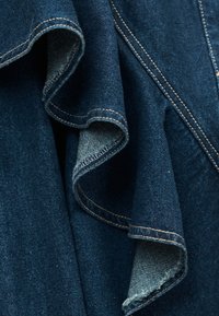 Tissu en denim bleu foncé avec surpiqûres marron visibles, montrant les bords repliés et le tissage texturé en gros plan.