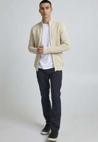 Lichtbeige ritsvest met geribbelde textuur, gecombineerd met een wit t-shirt en donkerblauwe broek. Zwarte sneakers maken de outfit compleet.