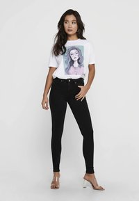 Biała koszulka z kolorowym nadrukiem kobiety, zestawiona z czarnymi, wysokim stanem i obcisłymi dżinsami oraz przezroczystymi sandałami na grubym obcasie.