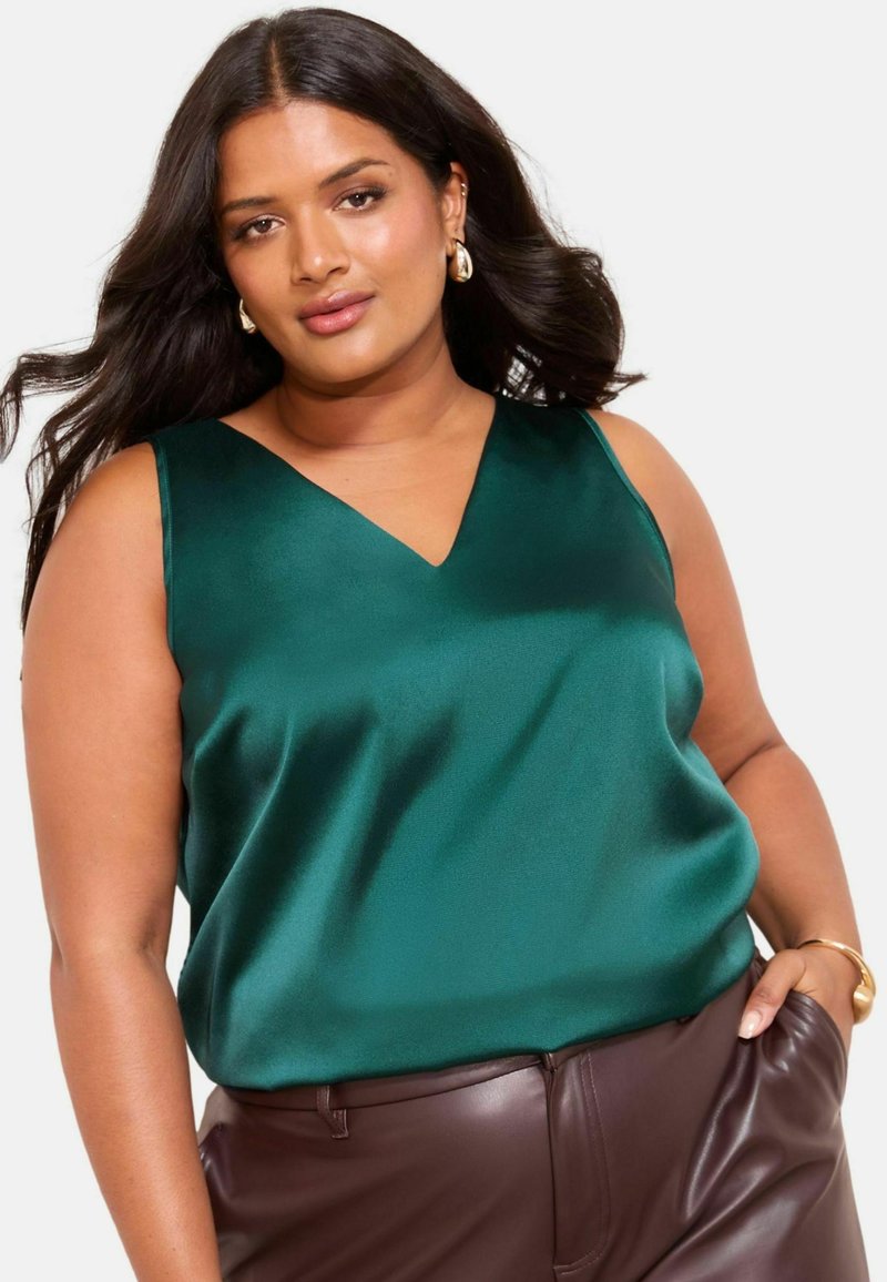 Top satin verde smeraldo senza maniche con design a V, texture liscia e vestibilità morbida, abbinato a pantaloni in pelle marrone.