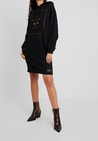 Kvinna i svart oversized hoodieklänning med guldlogo och bruna snokskinnsankelstövlar, stående mot en enkel ljus bakgrund.