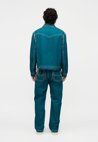 Άνδρας με σγουρά μαλλιά που φοράει τζάκετ και παντελόνι σε τιρκουάζ με αντίθετες λευκές ραφές, στέκεται με την πλάτη προς το φακό σε απλό φόντο.