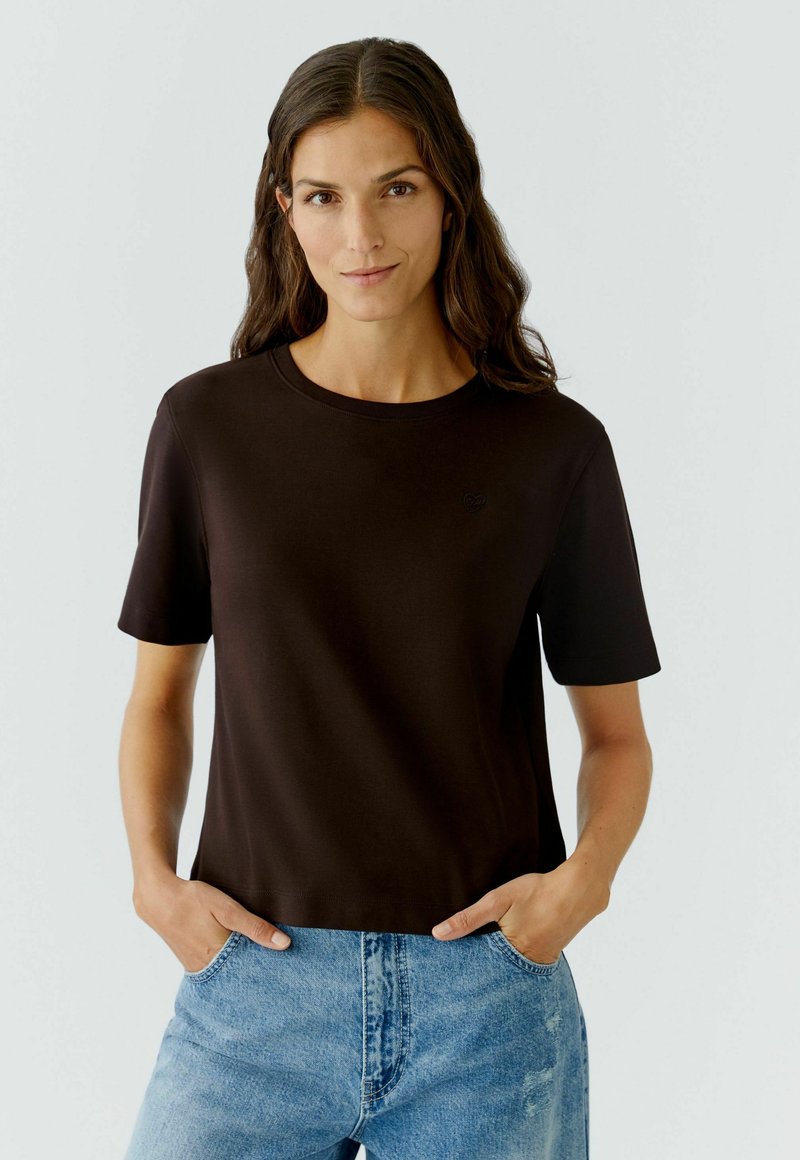 Bruine, t-shirt met korte mouwen van zacht materiaal, ronde halslijn, met een klein zwart logo op de borst. Gekozen om te dragen met blauwe jeans.