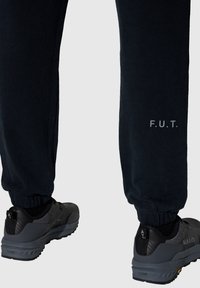 Schwarze Jogginghose mit elastischen Bündchen, mit dem Text "F.U.T." in hellgrau. Kombiniert mit schwarzen Sportschuhen. Weicher Stoff.