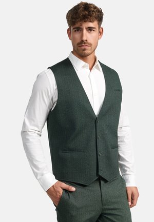 Chaleco de espiga verde hecho de tejido texturizado, con un cierre de cinco botones, dos bolsillos frontales y llevado sobre una camisa blanca.