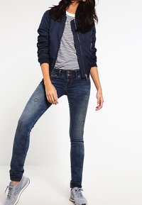 Námořnická bomber bunda přes pruhované tričko, doplněná o ošumělé modré skinny džíny a šedé tenisky. Ležérní a přiléhavý design.