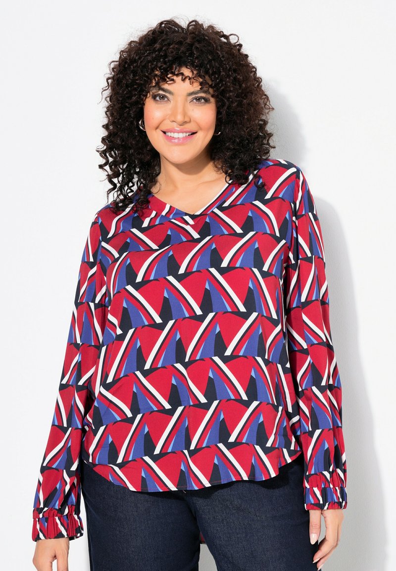 Langärmlige Bluse mit einem geometrischen Muster in Rot, Blau, Weiß und Schwarz, V-Ausschnitt und gerafften Manschetten. Glatte Stofftextur.