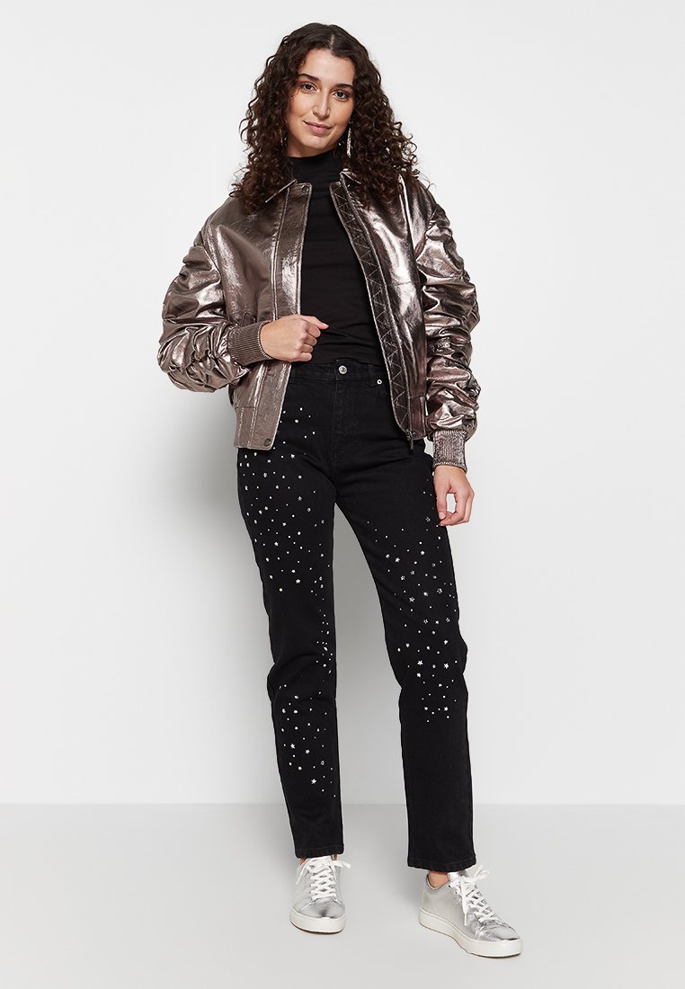 Metallische Bomberjacke in Roségold, kombiniert mit schwarzen Jeans, die mit sternartigen Verzierungen versehen sind. Das Outfit wird durch silberne Sneaker ergänzt.