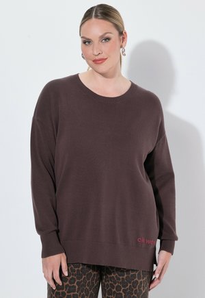 Vrouw draagt een losse bruine trui met geribbelde boorden en zoom, gecombineerd met een broek met luipaardprint, staand tegen een effen witte achtergrond.