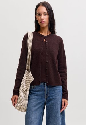 Femme aux longs cheveux foncés portant un cardigan boutonné marron foncé, un jean bleu et un sac cabas beige à carreaux sur l'épaule.