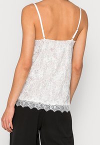 Žena oblečená v bílé krajkou olemované tílko s nastavitelnými ramínky a černými kalhotami, viděná zezadu na jednolitém pozadí.