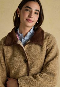 Kohvane fliistekk, millel on tekstuuriga pind, tumedast pruunist korduurist krae, nööpidega kinnitamine ja mugav lõige. All on sinine volangpaita.