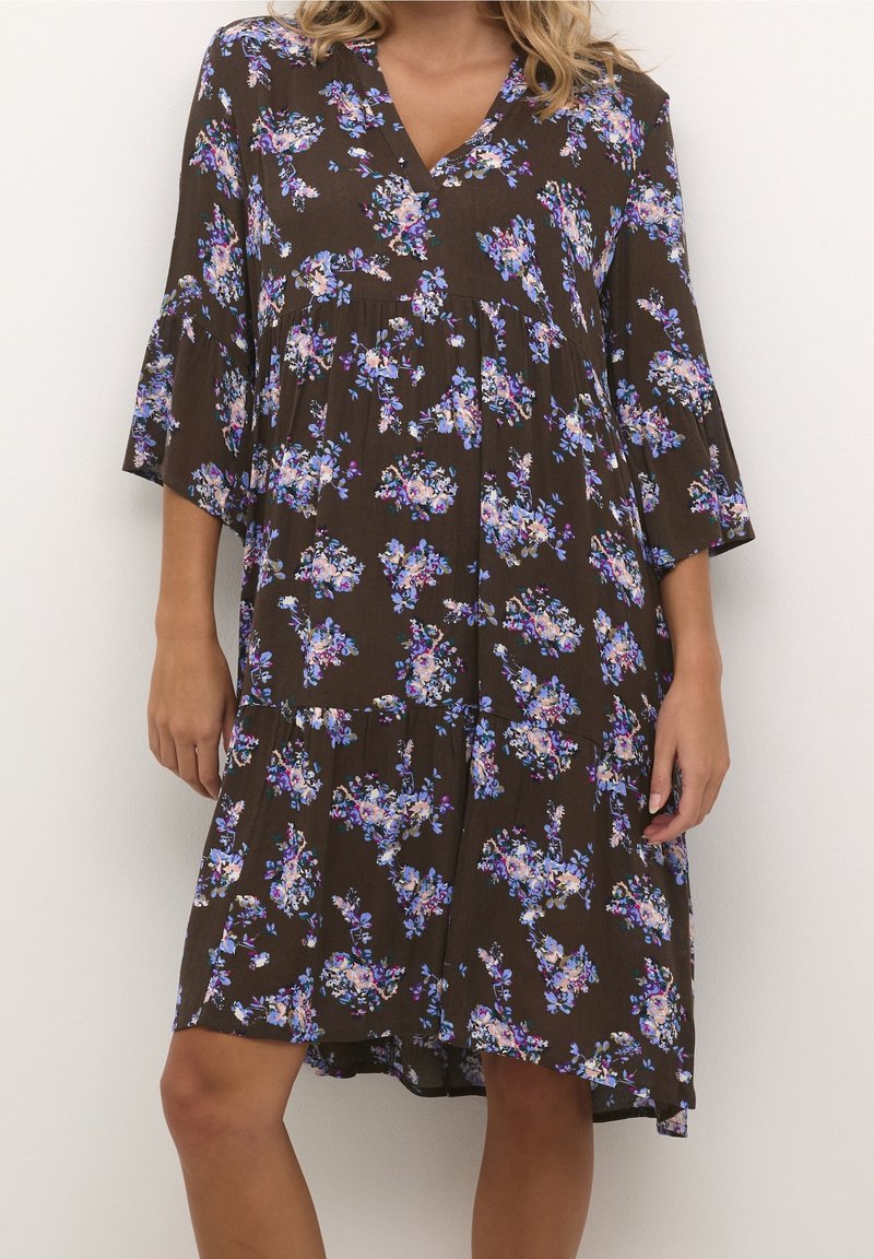 Femme portant une robe marron ample avec un imprimé floral violet et blanc et des manches trois-quarts, debout devant un fond uni.