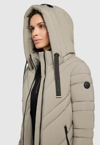 Giacca a vento beige chiaro con colletto alto, grande cappuccio, texture trapuntata orizzontale, due tasche laterali con zip e dettagli in metallo nero.