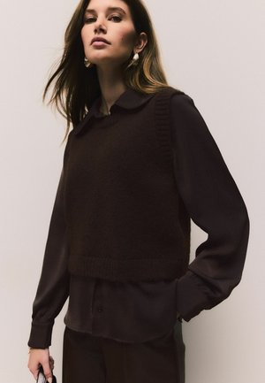 Brauner gestrickter Weste über einem dunklen Langarmshirt mit Kragen und geripptem Saum. Das Model trägt auffällige Ohrringe und hält eine Clutch.