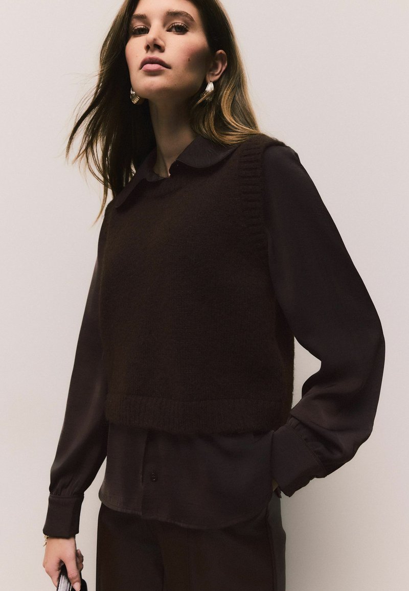 Brown knitted vest over a dark long-sleeve shirt, featuring a collar and ribbed hem. Model wears statement earrings and holds a clutch.