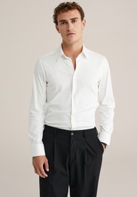 Camicia bianca a maniche lunghe con colletto classico e chiusura a bottoni, abbinata a pantaloni neri plissettati; tessuto liscio e design aderente.