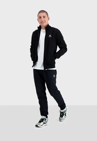 Giacca nera con zip sopra una t-shirt bianca, abbinata a pantaloni jogger neri e sneaker sportive nere e bianche. Presenta dettagli del logo.