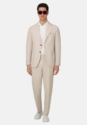 Hombre vestido con traje beige, camisa blanca, zapatillas blancas, pañuelo en el bolsillo y gafas de sol oscuras, de pie frente a un fondo claro.