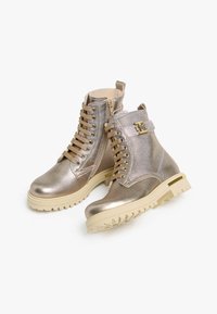 Metallisch goldene Knöchelstiefel mit strukturiertem Finish, ausgestattet mit beigen Schnürsenkeln, einem seitlichen Reißverschluss und einer klobigen, hell gefärbten Sohle. Goldfarbene Beschläge setzen Akzente.