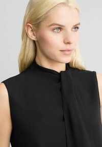 Zwarte mouwloze blouse met een hoge kraag en een strikdetail aan de voorkant. Glad stof met een zachte textuur, aansluitend ontwerp en geen zichtbare patronen.