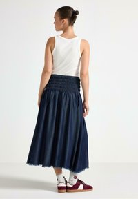 Denim maxi nederdel med smock-detalje i taljen, frynset kant, navy farve. Båret med en hvid tanktop og burgunder sneakers med hvide detaljer.