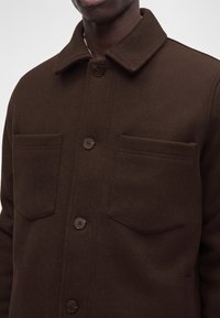 Veste en laine brun foncé avec fermeture à boutons et deux poches poitrine portée par une personne, cadrée sous le nez et au-dessus de la taille.
