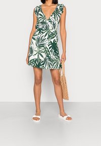 Robe florale verte et blanche avec des manches volantes, décolleté en V profond et taille cintrée. Accompagnée de sandales blanches et d'un sac en osier texturé.