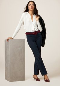 Witte blouse met lange mouwen, marineblauwe broek met een bordeauxrode riem, en donkerrode puntige hakken. Betonnen blok dient als display voet.