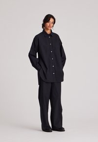 Camicia nera oversize con colletto e taschino anteriore, caratterizzata da chiusure a bottone. Abbinata a pantaloni neri a gamba larga e scarpe nere.