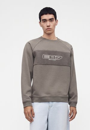 Giovane uomo che indossa una felpa grigia a maniche lunghe con pannello frontale strutturato e logo "EA7 Emporio Armani", abbinata a jeans azzurri.