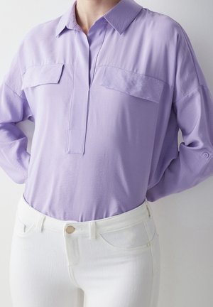 Personne portant un chemisier lavande à manches longues rentré dans un pantalon blanc taille haute, debout les mains sur les hanches devant un fond uni.