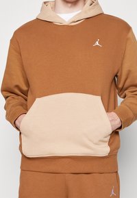 Homme portant un sweat à capuche marron et beige avec poche kangourou et logo Jordan blanc sur la poitrine, accompagné d'un pantalon marron assorti avec logo.