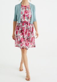 Květinové šaty v odstínech růžové a zelené s nařaseným výstřihem a zavazováním v pase, doplněné světle modrým krátkým kardiganem s krátkým rukávem.