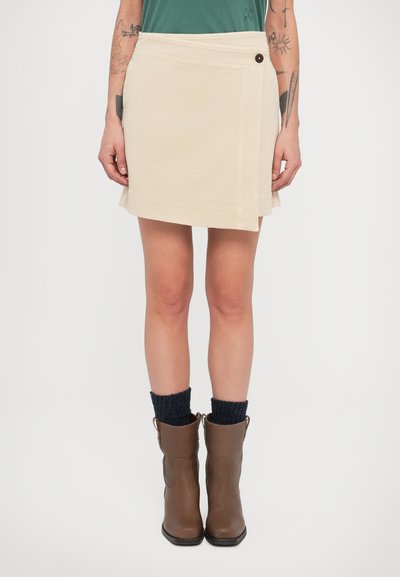 Jupe portefeuille beige avec fermeture par bouton, coupe droite et surpiqûres contrastées. Portée avec des bottes marron et des chaussettes en maille foncées.