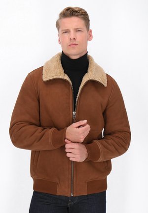 Braune Wildleder-Bomberjacke mit einem hellbraunen Kunstpelzkragen, Frontreißverschluss und gerippten Bündchen. Das Model trägt einen schwarzen Rollkragenpullover darunter.