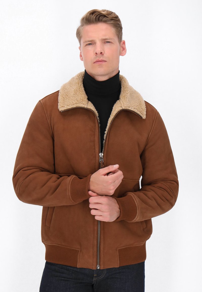 Brun suede bomberjakke med en beige faux fur-krave, front lynlås og ribbede manchetter. Modellen har en sort turtleneck på nedenunder.