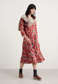 Femeie îmbrăcată cu o rochie midi roșie cu flori și mâneci 3/4, sosete gri, saboți maro și un pulover bej aruncat pe umeri, pe un fundal alb.