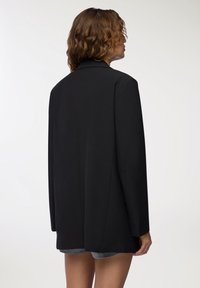 Blazer nero con una vestibilità su misura, caratterizzato da un colletto appuntito e maniche lunghe. Il tessuto è liscio e strutturato, con un taglio di lunghezza media.