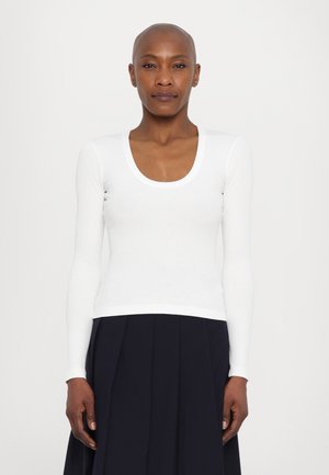 Ribbed white long-sleeve top with a scoop neckline, fitted shape, and stretchy fabric, worn with a pleated navy skirt.