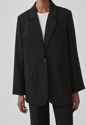 Persoon die een zwarte blazer met één knoop draagt over een wit topje, met bijpassende zwarte broek, staand tegen een effen achtergrond.