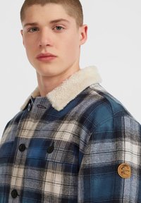 Young man wearing a blue and white plaid jacket with black buttons and a white fleece collar, looking slightly to the side.