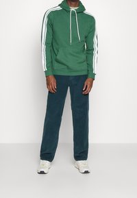 Sudadera verde con rayas blancas en las mangas, bolsillo frontal y cordones. Combinada con pantalones de pana oscuros y zapatillas deportivas.