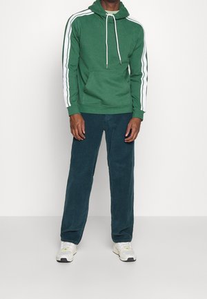 Groene hoodie met witte strepen op de mouwen, een voorkantzak en een trekkoord. Gecombineerd met donkere corduroypantalons en sportschoenen.
