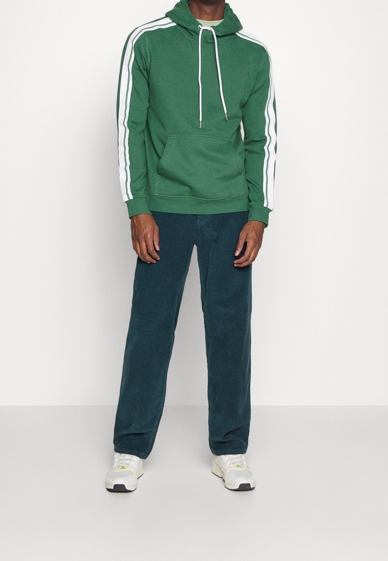 Sudadera verde con rayas blancas en las mangas, bolsillo frontal y cordones. Combinada con pantalones de pana oscuros y zapatillas deportivas.