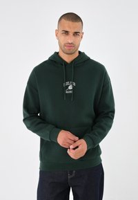 Hanorac verde închis cu buzunar frontal, glugă cu șnur și un logo alb care spune "Fidel Club Madrid". Material moale și croială lejeră.
