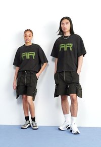 T-shirts pretos oversized com gráfico verde "AR", combinados com calções cargo pretos que apresentam vários bolsos e fechos éclair. Calçado casual.