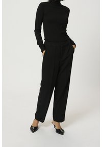 Un pull à col roulé noir associé à un pantalon noir taille haute. Le pantalon présente une coupe droite et des poches latérales. Des chaussures noires à talons hauts avec des pointes.