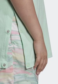 Světle zelená bavlněná košile s odnímatelnými sklapovacími knoflíky, která se vyznačuje hladkou strukturou. Pod ní je viditelná multikolorová látka s vlnitým vzorem.
